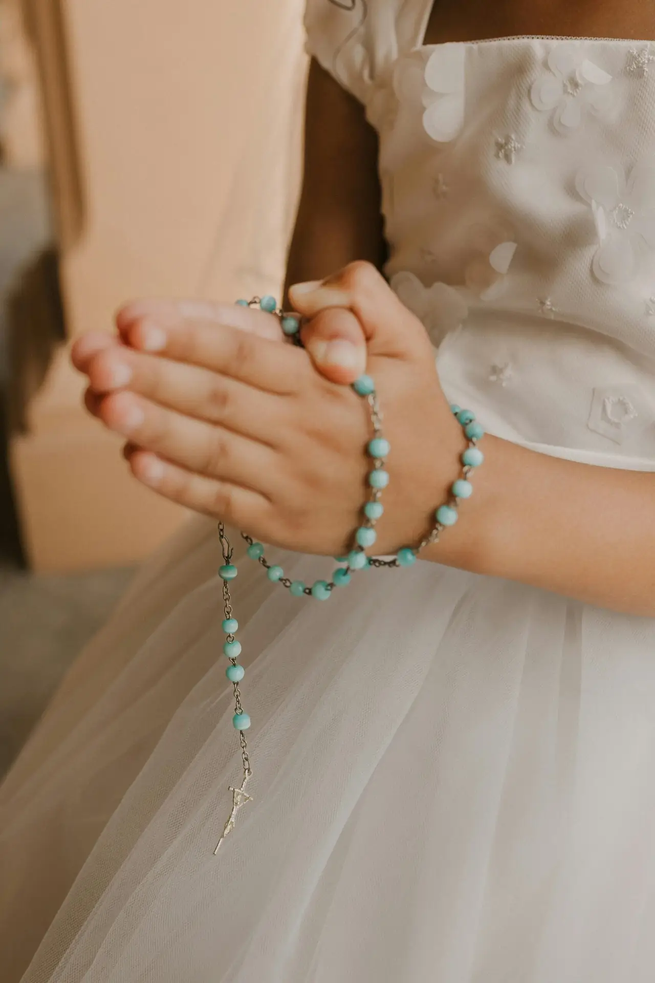 A close-up of a child praying with turquoise rosary beads in a white dress.